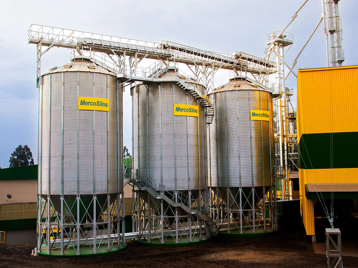 Tres silos MercoSilos de fundo conico sobre estrutura metalica, com escada de acesso e barracao amarelo ao fundo