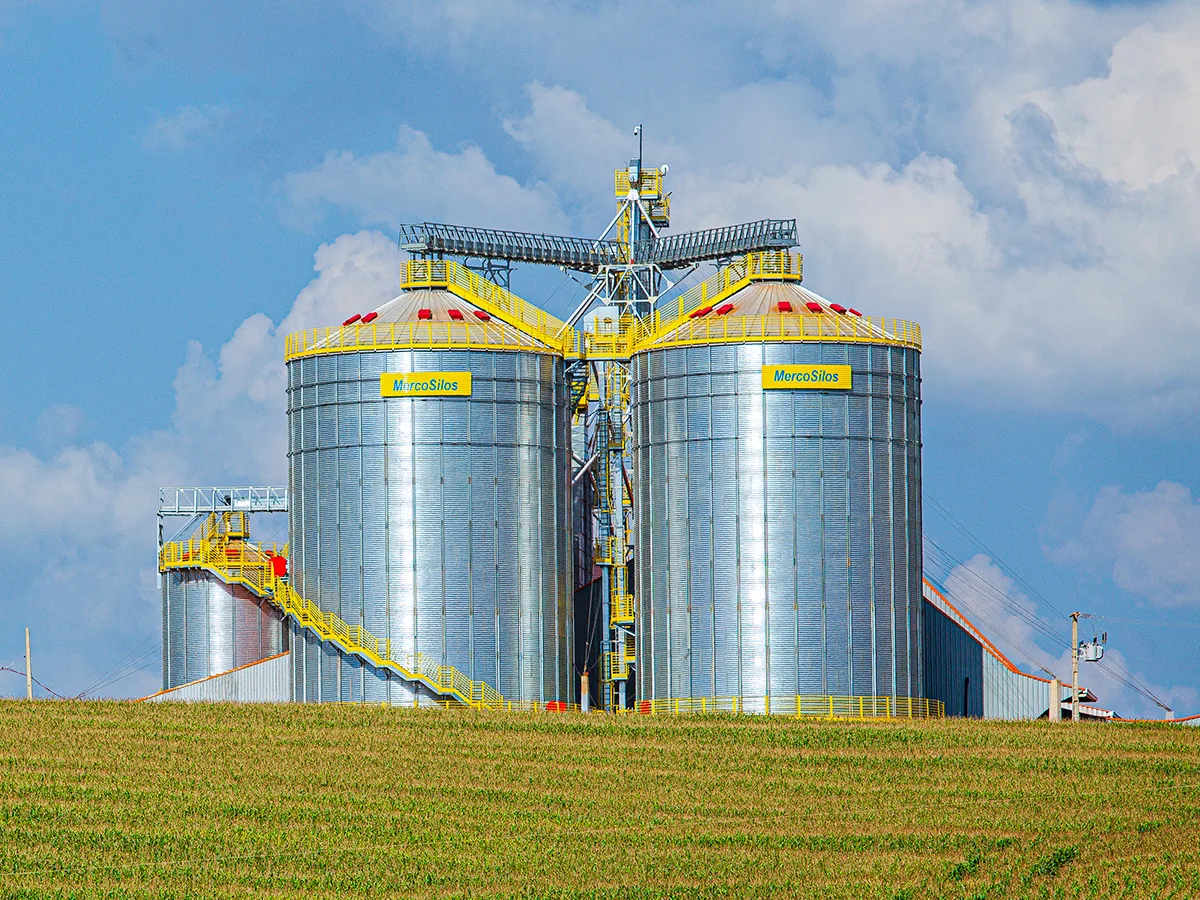 Dois silos MercoSilos de fundo plano com telhado conico, passarela superior e escada lateral, instalados ao lado de um barracao