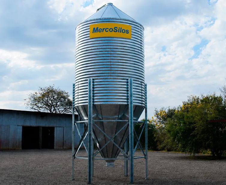 Silo granjeiro MercoSilos em chapa galvanizada corrugada com fundo conico e estrutura de sustentacao, ao lado de um galpao