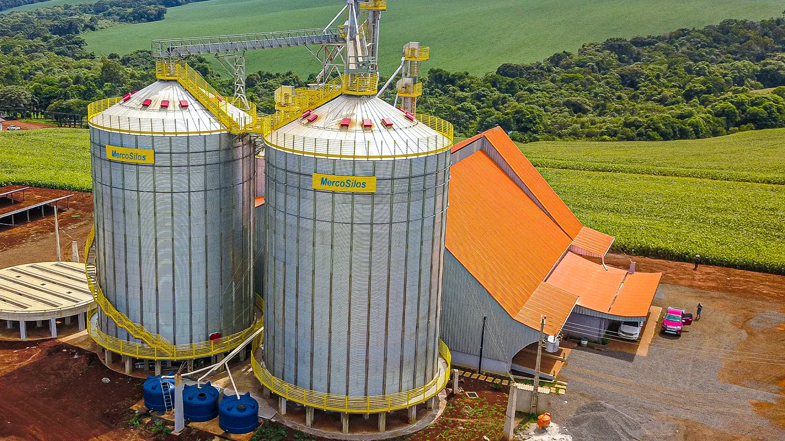 Bateria de silos MercoSilos de fundo plano em area rural, com elevador de cereais e passarela metalica ligando os topos